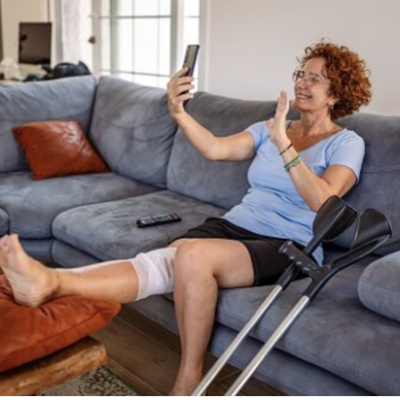 A woman with a bandage on her knee and crutches close by. She is on her sofa, video chatting on her phone. 